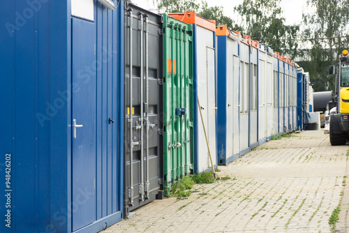Row of industrial storage containers and portable site offices aligned along a paved walkway at a construction or logistics area, showing modular buildings and container units used for on-site work 