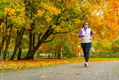 Pretty middle-aged blonde woman dressed sporty running on background of beautiful autumnal trees in city park on autumn day. Front view. Autumnal training. Healthy running.