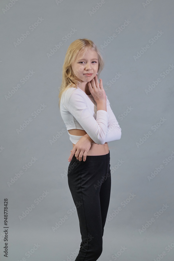 Dressed in a white crop top and black pants, a girl with blonde hair poses with her hand near her face, exuding a thoughtful expression against a simple backdrop. This photo captures modern, youthful Dressed in a white crop top and black pants, a girl with blonde hair poses with her hand near her face, exuding a thoughtful expression against a simple backdrop. This photo captures modern, youthful