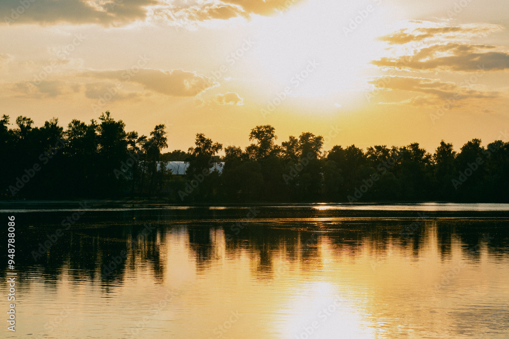 Fototapeta premium A bright light orange sunset on a calm river with a bank lined with tall trees. The rays of the luminary glare and reflect from the water surface. Sunny evening in nature.