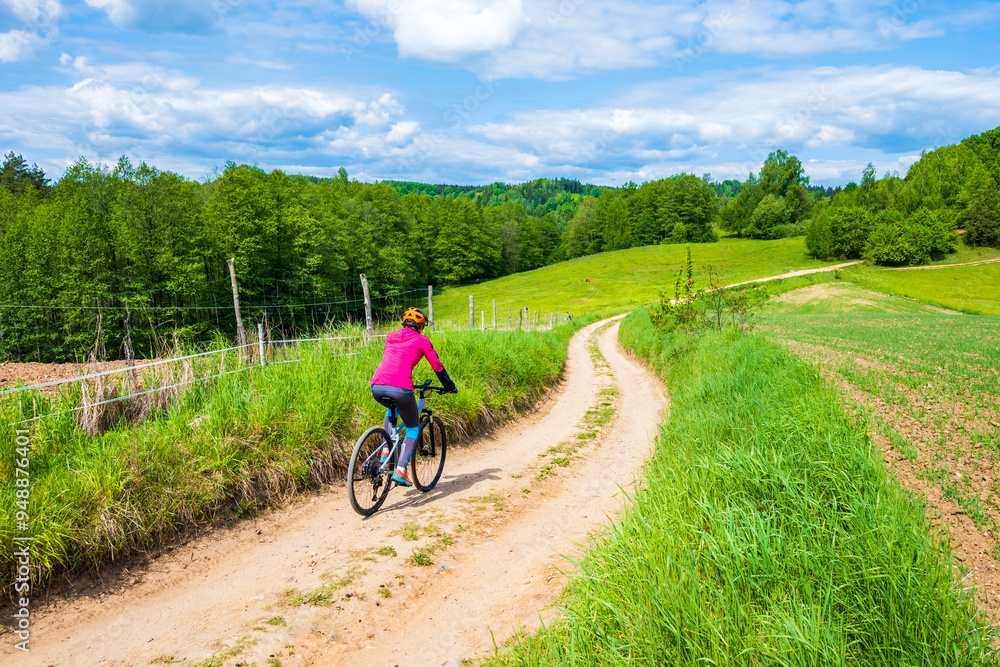 Woman cyclist riding bicycle on gravel countryside road along green meadows and hills, Suwalski Landscape Park, Podlasie, Poland
