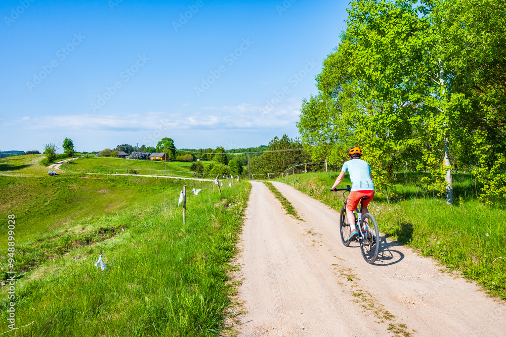 Woman cyclist riding bicycle on gravel rural road among green meadows and fields, Suwalski Landscape Park, Podlasie, Poland