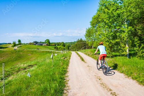Obraz na płótnie Woman cyclist riding bicycle on gravel rural road among green meadows and fields, Suwalski Landscape Park, Podlasie, Poland w salonie