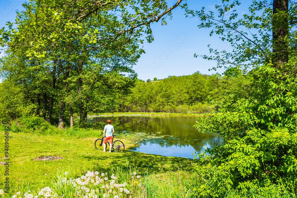 Woman cyclist standing on lake shore beach and among green meadows, Suwalski Landscape Park, Podlasie, Poland