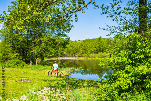 Obraz na płótnie Woman cyclist standing on lake shore beach and among green meadows, Suwalski Landscape Park, Podlasie, Poland w salonie