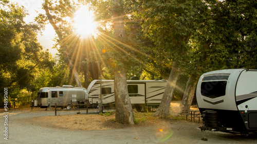 Rv camping trailers parked at campsite under trees with sunburst coming through 