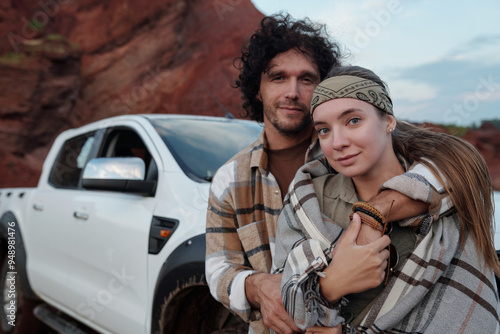 Happy young man in flannel embracing his pretty girlfriend wrapped into grey checkered plaid while both looking at camera