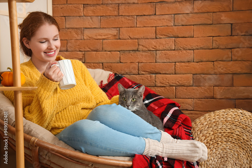 Young woman with cup of tea and cute cat sitting in armchair at home