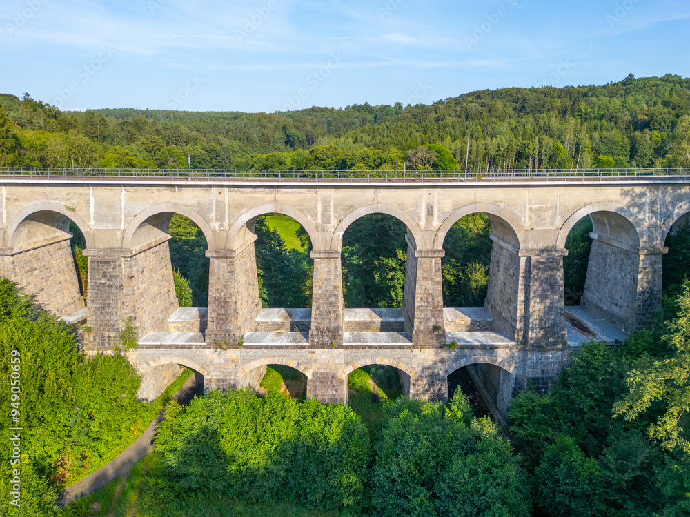 Fototapeta premium The Sychrov Railway Bridge rises gracefully above lush greenery, showcasing its impressive arches against a backdrop of vibrant trees.