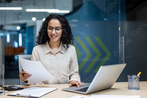 Smiling businesswoman reviewing documents at work desk. She uses laptop while analyzing paperwork, showcasing professionalism and engagement in modern office setting.