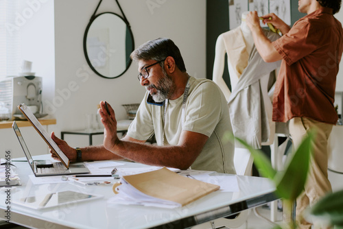 Indian professional designer discussing details with customer via smartphone while tailor creating new dress photo