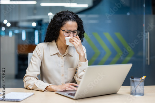 Woman in office setting working on laptop, holding tissue, showing signs of being unwell. She wears glasses and focuses on her work, reflecting concept of managing health in professional environment. photo