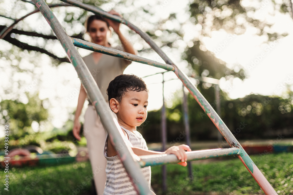 An Asian boy is playing on monkey bars in a playground while his mother stands nearby to help and ensure his safety. Active play and the joy of developing skills. An Asian boy is playing on monkey bars in a playground while his mother stands nearby to help and ensure his safety. Active play and the joy of developing skills.