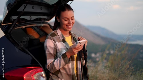 Beautiful, young woman getting ready for hiking on the mountain and using her smartphone while leaning on the car trunk