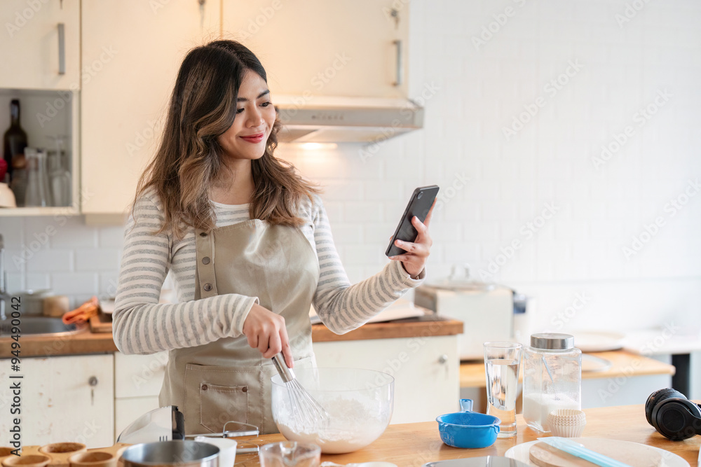 Smiling young Asian woman checking phone while mixing batter Smiling young Asian woman checking phone while mixing batter