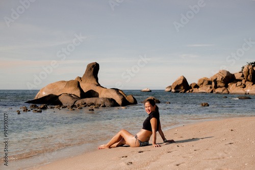 View of Asian woman in black bikini swimsuit sitting on the beach on sunny day, enjoying sunshine. Summer, holiday, travelling, vacation. Woman relax by ocean for summer vacation. Samui, Thailand.