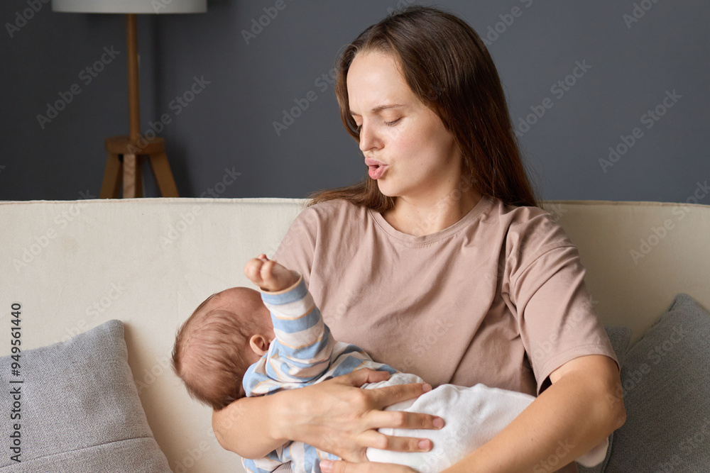 Caucasian woman wearing casual clothing mommy rocking her newborn baby while sitting on sofa in living room holding infant son or daughter trying to put the child to bed saying shh Caucasian woman wearing casual clothing mommy rocking her newborn baby while sitting on sofa in living room holding infant son or daughter trying to put the child to bed saying shh