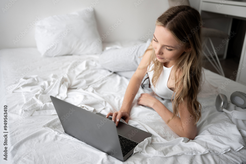 Young attractive woman lying in bed at home and using laptop computer. Female student looking at computer Young attractive woman lying in bed at home and using laptop computer. Female student looking at computer