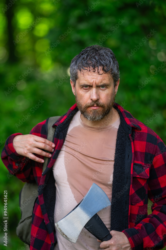 Fototapeta premium Confident lumberjack works in summer forest. Man with an ax. Woodcutter in plaid shirt in forest. Hipster with axe. Lumberjack with an axe. Bearded lumberjack. American lumberjack with hatchet.