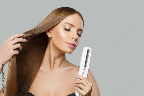 Cheerful fashion model with brown shiny straight haistyle portrait. Cute woman with straightening iron in her hand on gray studio background photo