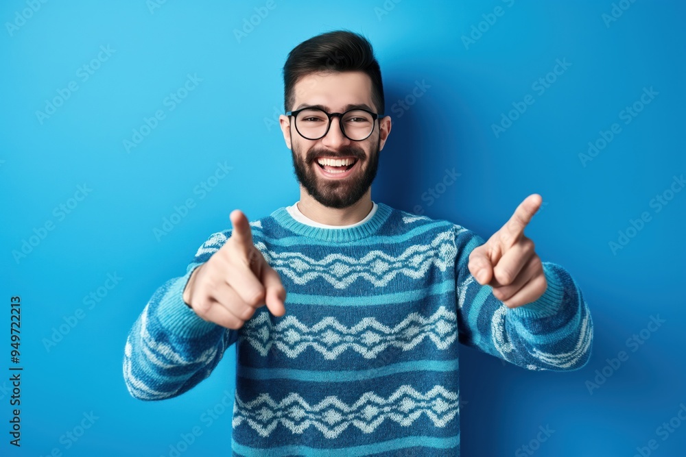 Naklejka premium Handsome bearded man in casual sweater and glasses standing in front of blue background. He smiling and pointing to the camera with fingers, giving a thumbs up.