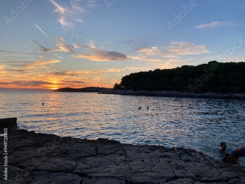Sunset in Croatia, Pula. Amazing view of the orange sunset with blue sky and clouds on the sea and some trees in the forest in the background with rocky beach. 