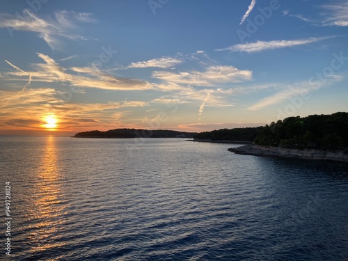 Sunset in Croatia, Pula. Amazing view of the orange sunset with blue sky and clouds on the sea and some trees in the forest in the background with rocky beach. 
