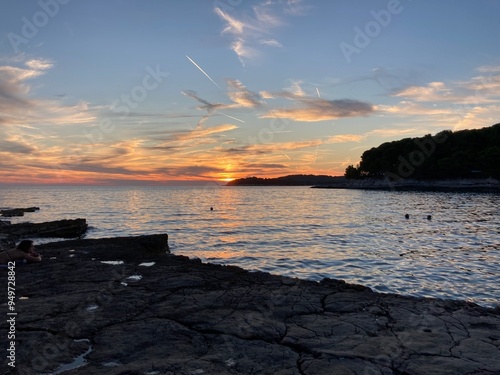 Sunset in Croatia, Pula. Amazing view of the orange sunset with blue sky and clouds on the sea and some trees in the forest in the background with rocky beach. 