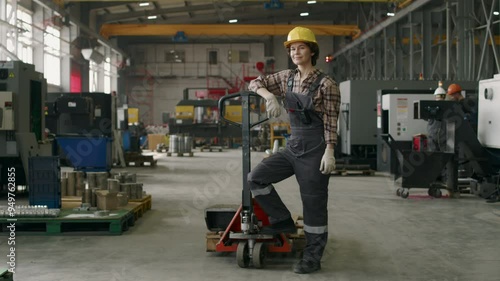 Long shot of confident female factory worker wearing uniform with safety helmet posing for camera with pallet jack