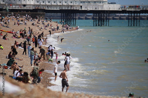 Brighton beach, East Sussex, UK - 08.08.2024: Brighton beach people sunbathing on shingle pebble beach sunny day with blue sky, Brighton pier behind photo