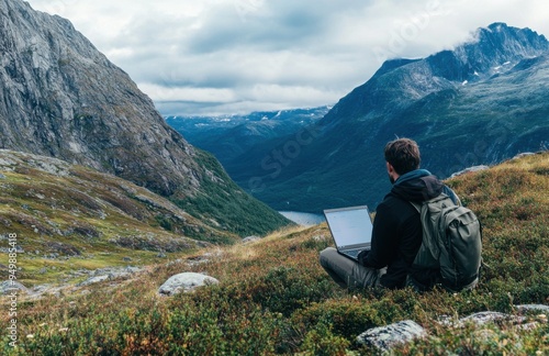 Man sits on the grassy ground with his laptop, surrounded by mountains and forests. He gazes at the magnificent scenery of fjords while working online or writing an academic paper