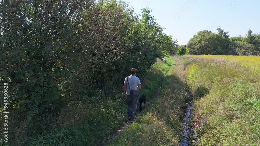 A man in light clothes and linen pants walks his dog along a path through wild meadows and trees, a bag over his shoulder, enjoying the sunny day. A man in light clothes and linen pants walks his dog along a path through wild meadows and trees, a bag over his shoulder, enjoying the sunny day.