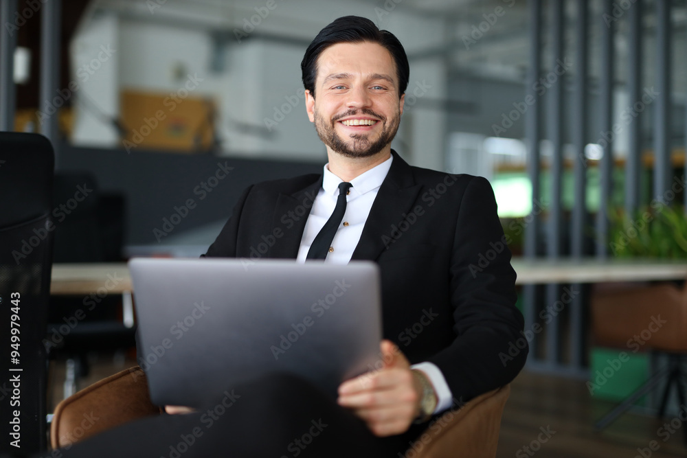 A man in a suit and tie is sitting in a chair with a laptop in front of him. He is smiling and he is enjoying himself A man in a suit and tie is sitting in a chair with a laptop in front of him. He is smiling and he is enjoying himself