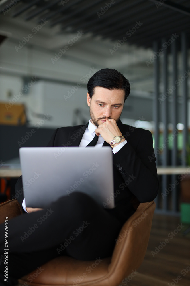 A man in a suit is sitting in a chair with a laptop open in front of him. He is deep in thought, possibly working on a project or problem. Concept of focus and concentration A man in a suit is sitting in a chair with a laptop open in front of him. He is deep in thought, possibly working on a project or problem. Concept of focus and concentration