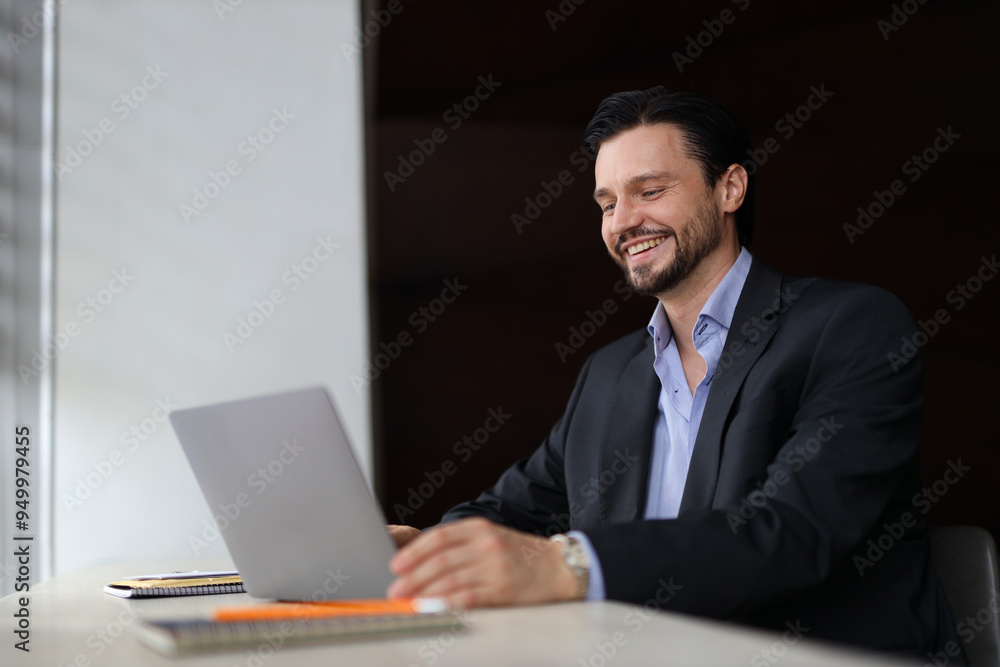 A man is sitting at a desk with a laptop open in front of him. He is smiling and he is enjoying himself A man is sitting at a desk with a laptop open in front of him. He is smiling and he is enjoying himself
