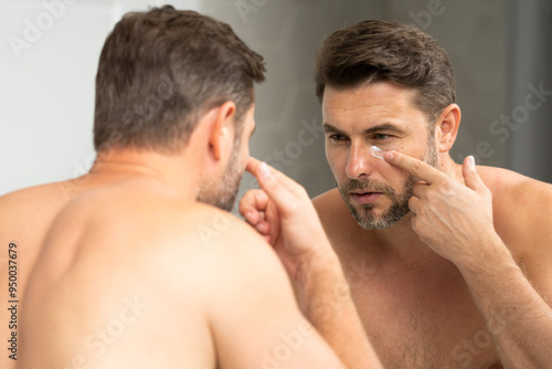 man in the bathroom moisturizing face cream. Happy young handsome man holding cream. Close up portrait of man 40s applying cream on face. Handsome serious man enjoy fresh perfect skin, applying cream.