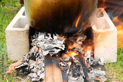  Large metal pot supported by cinder blocks over a wood fire, with flames and ash-covered logs visible beneath, set outdoors on grass. photo