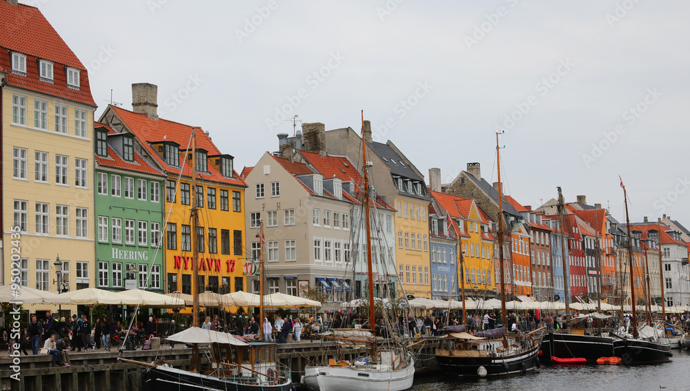 Naklejka premium Copenhagen, COP, Denmark - August 22, 2024: colorful houses in the canal called NYHAVN which means New Harbour with bars and restaurants and boats