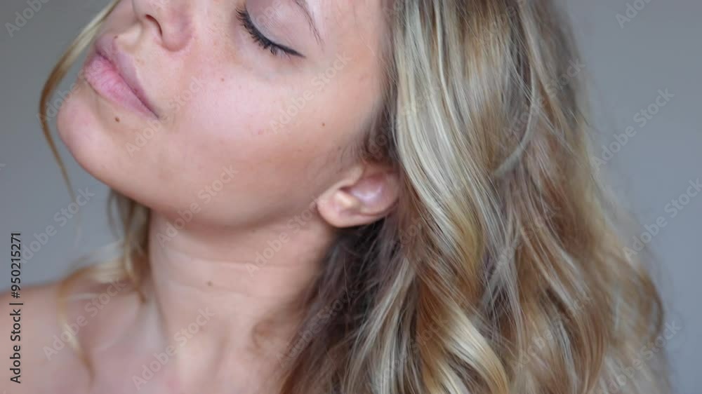 A young blonde woman showing her golden wavy hair touching locks with hand on a grey background. Result of coloring, highlighting, perming. Beauty and fashion concept