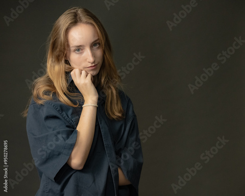 A young blonde woman in a loose blue shirt poses with her hand thoughtfully on her chin, creating a reflective and contemplative mood in a professional studio setting.