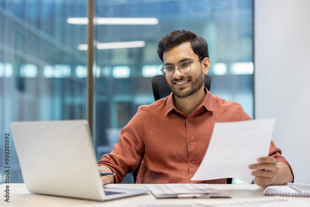 © Liubomir - Young professional man wearing glasses working happily at laptop in modern office. Holding document, showcasing productivity and success. Captures business environment, with sense of achievement