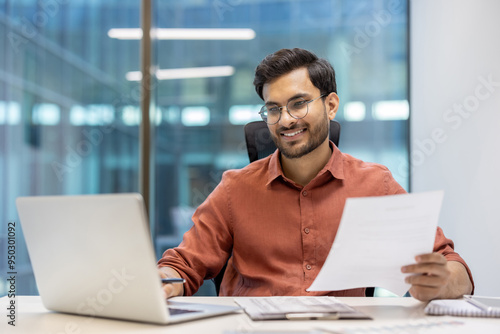 Young professional man wearing glasses working happily at laptop in modern office. Holding document, showcasing productivity and success. Captures business environment, with sense of achievement