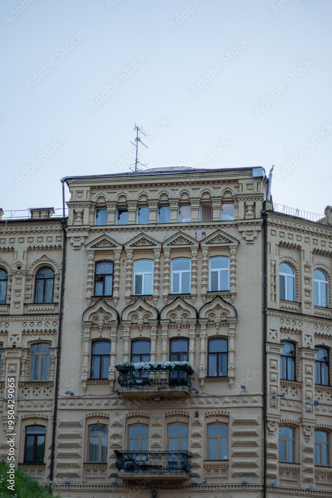 Obraz premium A detailed view of a historic, ornate apartment building with multiple floors, balconies adorned with flowers, and various window styles, against a clear blue sky. 