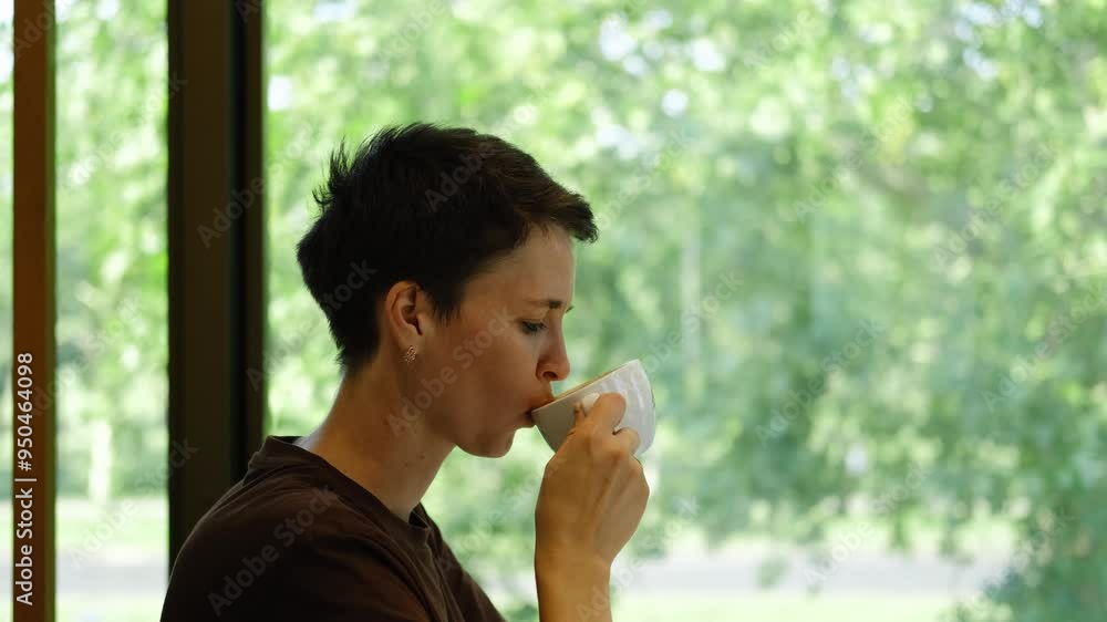A young handsome woman with short haircut is sitting in a cafe and drinking coffee from a white ceramic cup. The girl is sitting at a table by the window in a coffee shop and enjoying a break alone A young handsome woman with short haircut is sitting in a cafe and drinking coffee from a white ceramic cup. The girl is sitting at a table by the window in a coffee shop and enjoying a break alone