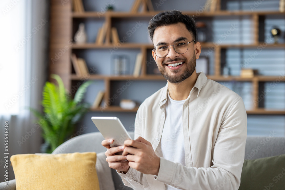 Smiling man holding tablet sitting on couch, enjoying digital content at home in modern living room setting Smiling man holding tablet sitting on couch, enjoying digital content at home in modern living room setting