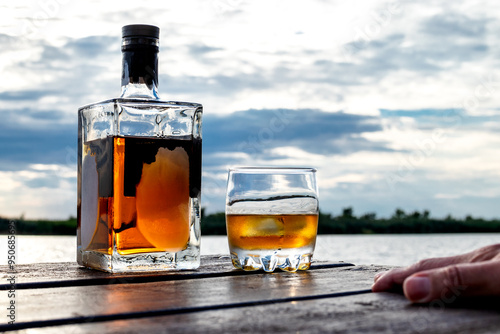 A chilled bottle and a glass of spirits on rocks are on a wooden dock by the water. Heavy clouds at sunset in the background.