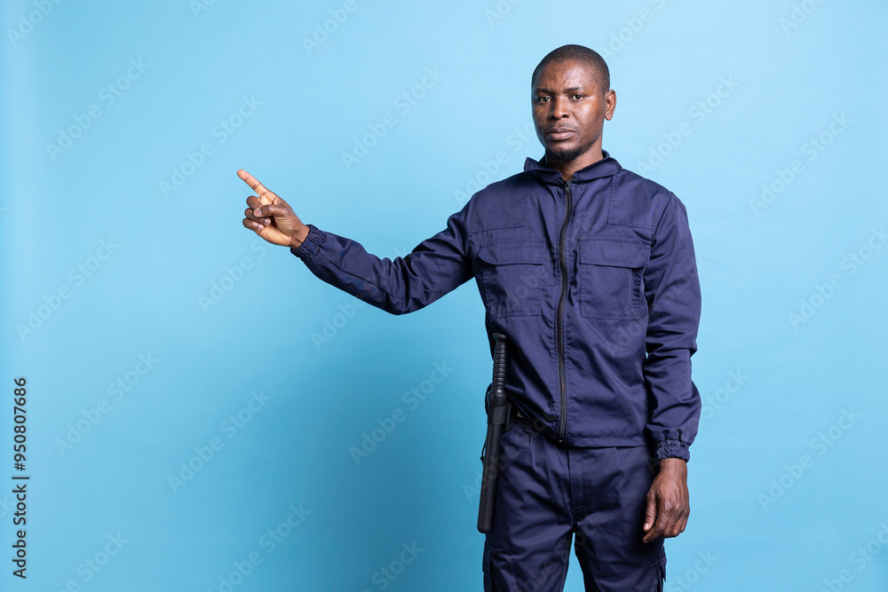 African american serious security agent in uniform pointing to the left side, creates a commercial ad to present something next to him. Guard raising his index finger, indicating a direction. African american serious security agent in uniform pointing to the left side, creates a commercial ad to present something next to him. Guard raising his index finger, indicating a direction.