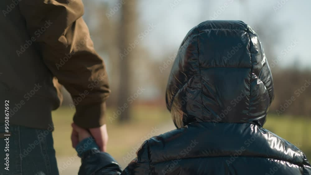 A close-up of a young boy holding hands with his father as they walk together, both dressed in warm jackets, with a blur view of trees in the background A close-up of a young boy holding hands with his father as they walk together, both dressed in warm jackets, with a blur view of trees in the background