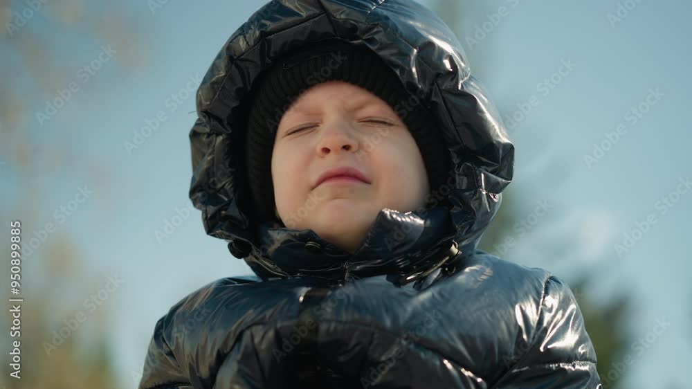 A close-up of a young child wearing a shiny black jacket, with a hood and a beanie, looking slightly frustrated and squinting as he open his eyes slowly A close-up of a young child wearing a shiny black jacket, with a hood and a beanie, looking slightly frustrated and squinting as he open his eyes slowly