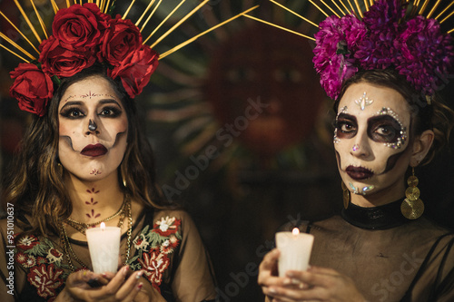 Mexican women dressed as Catrina with a headdress with flowers on their heads, alluding to the Day of the Dead, with a candle in their hands, under an arch of an old house. photo